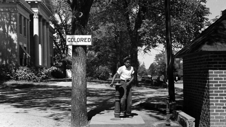 An African American child drinks from a water fountain by a sign labeled "colored."