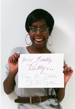 An African American woman holds a sign reading "you're really pretty...for a dark skin girl."