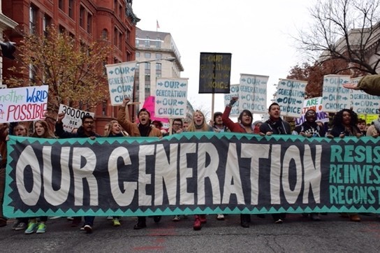 A diverse group of young people march behind a banner reading 'Our Generation.'