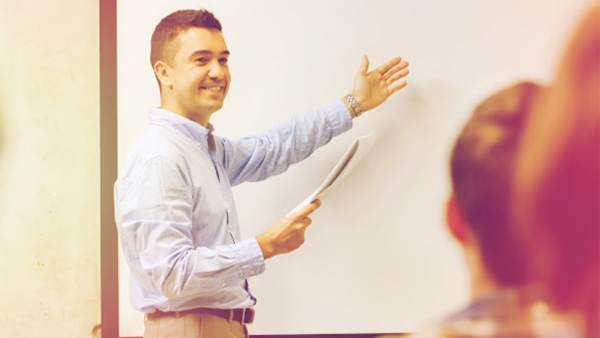 A person presents to an audience gesturing towards a whiteboard at the front of the room.